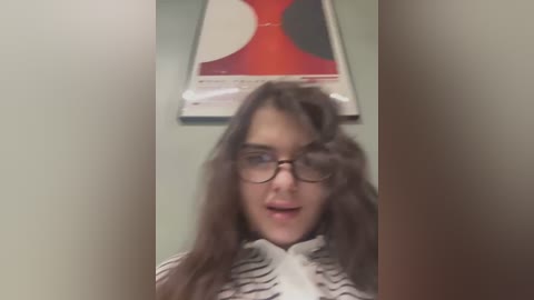 Media: A blurry video of a young woman with long brown hair, wearing glasses, and a striped shirt, standing in a room with a red and black abstract painting on the wall.