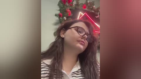 Media: A video of a young woman with long dark hair, wearing black-rimmed glasses and a striped polo shirt, standing in front of a Christmas tree with red ornaments and a glowing star.