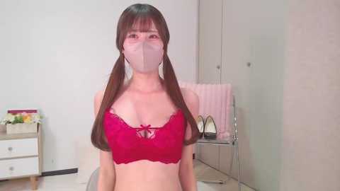 Media: Video of an Asian woman with long brown pigtails, wearing a red lace bra, face mask, and pink high heels, standing in a minimalist room with a white dresser and a chair.