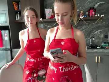 Media: Video of two young women with fair skin, wearing red aprons with \"Merry Christmas\" and snowflakes, standing in a modern kitchen with gray marble backsplash, dark cabinets, and festive decorations.