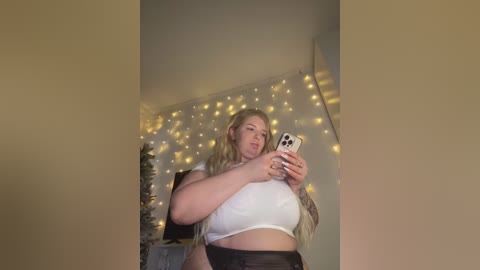 Media: Video of a curvy, light-skinned woman with long blonde hair, wearing a tight white crop top and black pants, taking a selfie in a dimly lit room with string lights and a Christmas tree in the background.