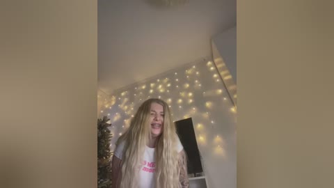 Media: A video of a blonde woman with long hair, wearing a white t-shirt with pink text, smiling, in a room with a Christmas tree, string lights, and a TV in the background.