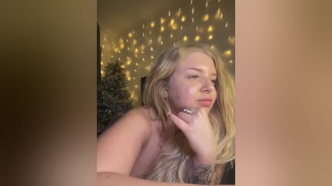 Media: Video of a topless, blonde woman with long hair, resting her chin on her hand, in a dimly lit room adorned with fairy lights and a decorated Christmas tree.