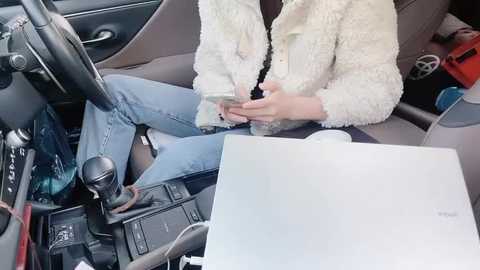 Media: Video of a person wearing a fluffy white coat, sitting in a car, texting on a smartphone, with a laptop open on the seat beside them.