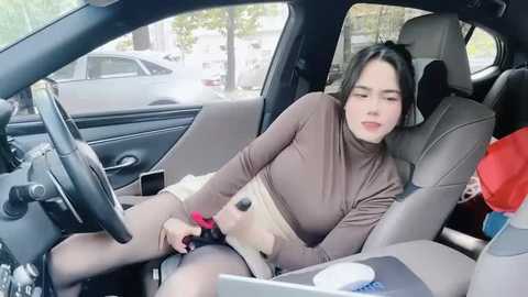 Media: Video of a young woman in a car, asleep with her head on the armrest, wearing a brown turtleneck and beige skirt. The car's interior is modern with gray leather seats.