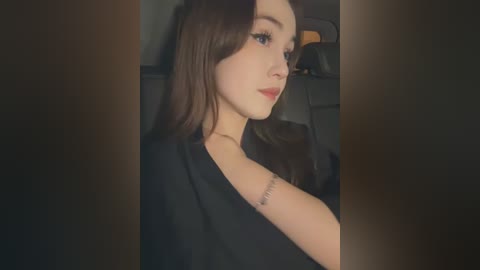 Media: Video of a young woman with fair skin, long brown hair, wearing a black top, sitting in a dimly lit car, with a tattoo on her left shoulder.