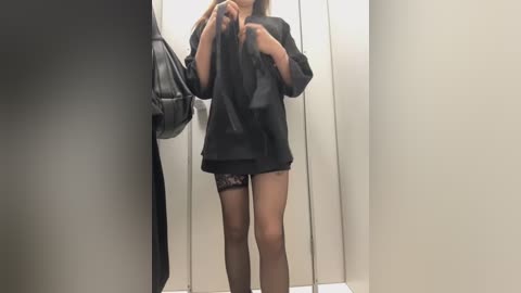 Media: A video of a slender, light-skinned woman in a black, see-through top and lace panties, holding a black handbag, standing in a narrow, beige-walled changing room.