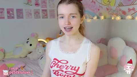 Media: Video of a young, slender, fair-skinned woman with light brown hair in pigtails, wearing a white \"Cherry Bomb\" tank top. She smiles, standing in a pastel-themed room with plush toys and fairy lights.