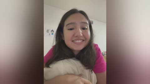 Media: Video of a young Asian woman with long dark hair, wearing a pink t-shirt, smiling and holding a white stuffed bear in a simple, brightly lit room with a white wall adorned with posters.