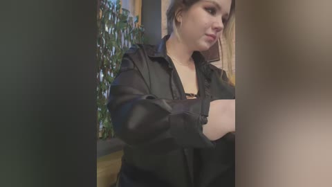 Media: Video of a pale-skinned, brunette woman with a medium build, wearing a black leather jacket, holding a black object, standing indoors with a green potted plant in the background.