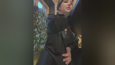 Media: Video of a woman in black leather outfit, grabbing a man's crotch, in a dimly lit office with a potted plant in the background.