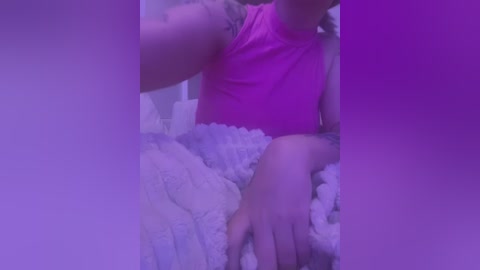 Media: A video of a person with fair skin, wearing a pink sleeveless top, partially covered by fluffy white bedding, taken in a dimly lit room with purple lighting.