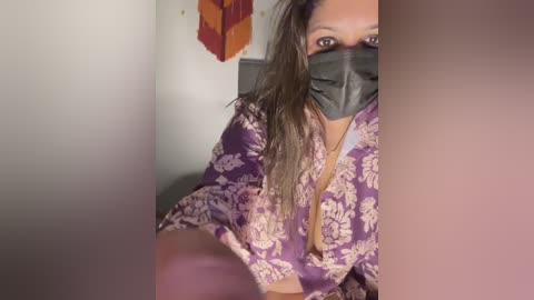 Media: Video of a South Asian woman with medium brown skin, wearing a purple floral-patterned silk robe, black mask, and black face shield, sitting indoors, partially obscured by a blurred foreground.