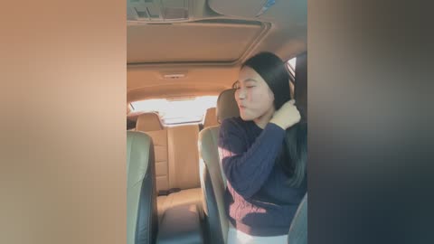 Media: Video of an Asian woman with long black hair, wearing a navy sweater, adjusting her seat belt in a beige leather car seat.