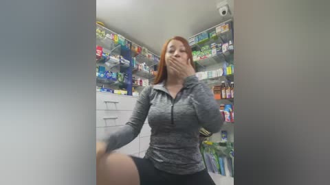 Video of a young woman with light skin and long red hair, wearing a gray athletic top and black shorts, covering her mouth with her hand, standing in a brightly lit pharmacy aisle filled with colorful products.