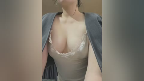 Media: Video of a light-skinned woman with large breasts, wearing a low-cut, gray blouse with lace trim, partially revealing cleavage. The background is blurred, with a hint of a chair and a neutral-colored wall.