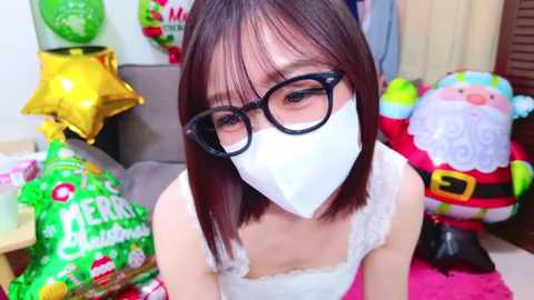 Media: A video of an Asian woman with straight, shoulder-length dark brown hair, wearing black-framed glasses, a white face mask, and a white lace tank top, in a festive room with Christmas decorations, including a Santa Claus figure and a green star.