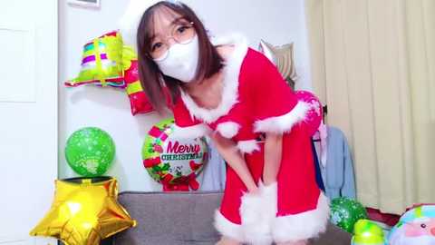 Media: Video of an Asian woman with glasses and a white face mask, dressed in a red Santa dress with white fur trim, bending over in a festive room with colorful decorations.