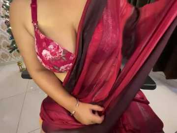 Media: A video of a woman in a red and maroon sari, revealing a pink floral bra. Her arm is wrapped around her waist, holding the sari. Background includes a patterned wall and tiled floor.