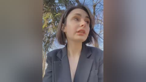 Media: Video of a young woman with short brown hair, wearing a dark blazer, standing outdoors in daylight, surrounded by bare trees and green foliage, looking contemplative.