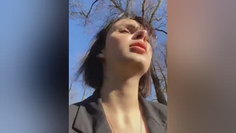 Media: Video of a fair-skinned woman with short dark hair, wearing a dark blazer, looking up in a park with bare trees and clear blue sky.