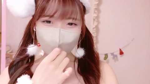 Media: Video of an Asian woman with long, straight brown hair and bangs, wearing a white face mask and fluffy, white Santa hat with red pom-poms. She holds a red ornament in her hand. Soft, pastel-colored background.