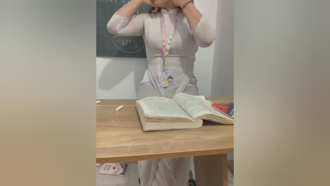 Media: Video of a woman in a light grey, form-fitting dress standing in a classroom with a wooden desk. She's adjusting her glasses, and an open book lies on the desk. The background features a chalkboard and a few scattered items on the floor.