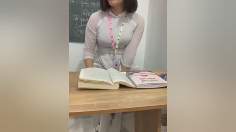 Media: Video of a woman with shoulder-length brown hair, wearing a light grey, semi-sheer blouse over a white top, seated at a wooden table. A lanyard with colorful beads hangs around her neck. An open book and a pink and white book with \"Honey\" on the cover are on the table. A chalkboard with scribbles is in the background.