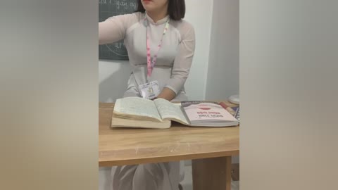 Media: Video of an Asian woman in a white sweater, seated at a wooden table with an open book and a pink lanyard. Background features a chalkboard with writing and a wooden chair.