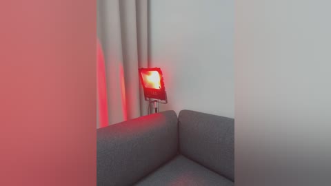 Media: Video of a modern, minimalist living room corner. A gray fabric couch with a textured surface is partially visible. A red LED light is mounted on the wall, casting a warm glow on beige curtains. The room is clean and simple, with a monochromatic color palette.