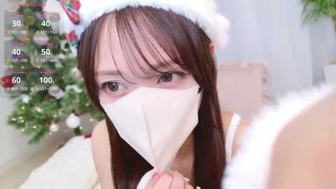 Video of an Asian woman with long brown hair, wearing a Santa hat, white mask, and a white top, holding a white cloth in front of her face. Background features a decorated Christmas tree.