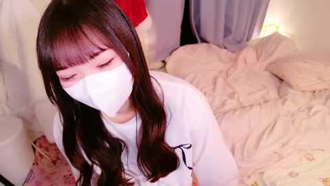 Media: Video of a young Asian woman with long, straight dark brown hair and blunt bangs, wearing a white face mask, sitting on a bed with rumpled white sheets and pillows.