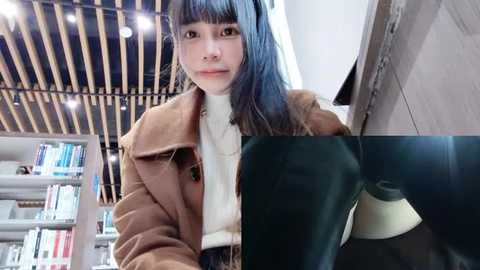 Media: Video of an East Asian woman with straight, dark blue hair and bangs, wearing a brown coat over a white turtleneck, sitting in a modern library with wooden ceiling beams, shelves filled with books, and a beige lampshade.