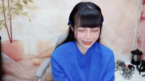 Media: A video of a young Asian woman with straight black hair, wearing a blue robe, smiling while seated in a room with a potted plant, a mirror, and glassware.