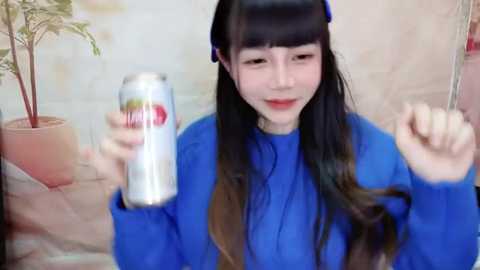 Media: Video of a smiling Asian woman with long black hair in a blue dress, holding a silver can and a potted plant in a beige room.