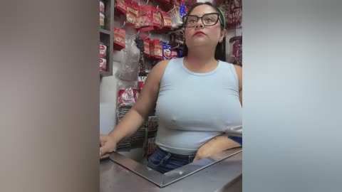 Media: A video of a plus-size Asian woman with glasses and dark hair, wearing a light blue tank top and jeans, working behind a cash register in a convenience store.