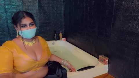 Media: A video of a woman with a curvy figure, medium skin tone, and large breasts, wearing a yellow blouse, face mask, and gold jewelry, sitting in a bathtub in a dimly lit, dark tiled bathroom.