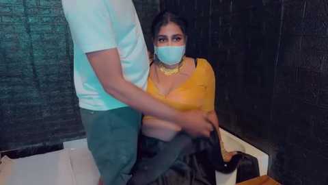 Media: Video of a South Asian woman with dark hair, wearing a yellow blouse and mask, seated on a wheelchair, being assisted by a man in a white shirt and green pants.