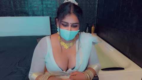 Media: A video of a South Asian woman with medium skin tone, wearing a white saree with a plunging neckline, blue face mask, and gold jewelry, sitting in a modern bathroom with a black tiled wall.