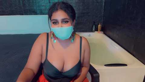 Media: Video of a plus-size woman with dark hair and dark skin, wearing a teal mask, black bra, and red skirt, seated on a dark tiled floor, in a modern bathroom with a white bathtub and black tiled wall.