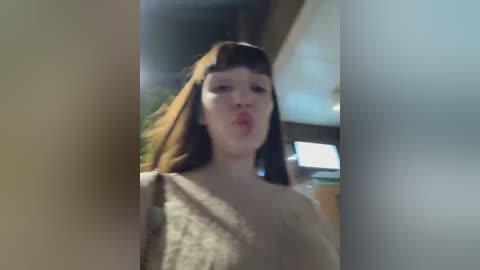 Media: A video of a young woman with fair skin, straight brown hair, and blunt bangs, making a pouty face. She wears a beige, sleeveless top. The background is blurry, showing an indoor setting with wooden beams and soft lighting.