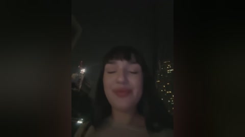 Media: Video of a woman with straight black hair and bangs, smiling with closed eyes, in a dimly lit room with a cityscape visible through a window.