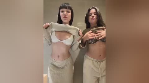 Media: Video of two women indoors, one pulling up a beige sweatshirt to reveal a white bra, the other lifting a brown crop top to expose a leopard print bra, both wearing matching beige sweatpants.