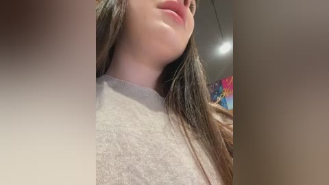 Media: Video of a young woman with fair skin, long brown hair, and full lips, wearing a light grey shirt, taken from a low angle, focusing on her face and neck. Background features colorful, abstract art.