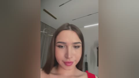 Media: A video of a young woman with fair skin, straight brown hair, and a red lipstick smile. She's framed by two vertical objects in a modern, well-lit room with a white ceiling and grey walls.