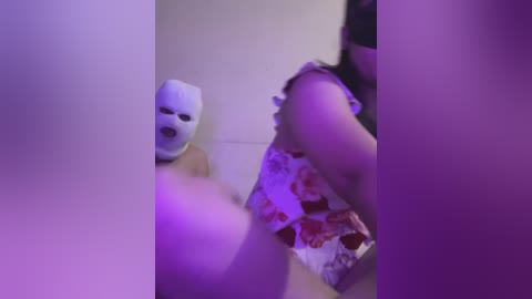 Media: Video of a woman in a white floral dress and a white mask, possibly a Halloween or cosplay outfit, taken in a dimly lit room with purple lighting, creating a mysterious atmosphere.