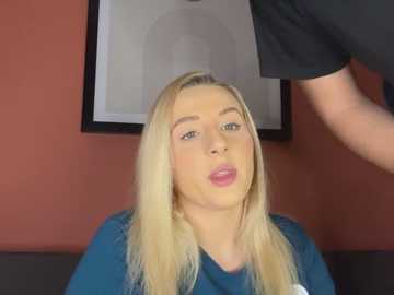 Media: Video of a blonde woman with fair skin, wearing a blue shirt, sitting indoors with a red wall and framed picture in the background.