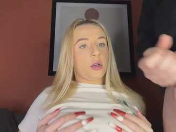 Video of a young blonde woman with fair skin and pink lipstick, lifting her white shirt, revealing her breasts. She's indoors with a red wall and framed art behind her.