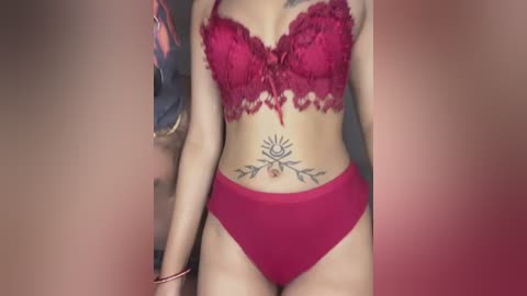 Media: Video of a light-skinned woman in a red lace bra and matching panties, showcasing a sun tattoo on her lower abdomen. Background is blurred.