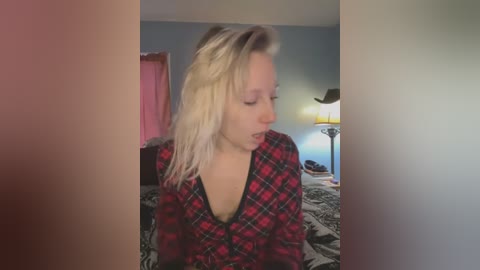 Media: Video of a blonde woman with light skin, wearing a red and black plaid shirt, sitting on a bed with a patterned duvet in a dimly lit room.